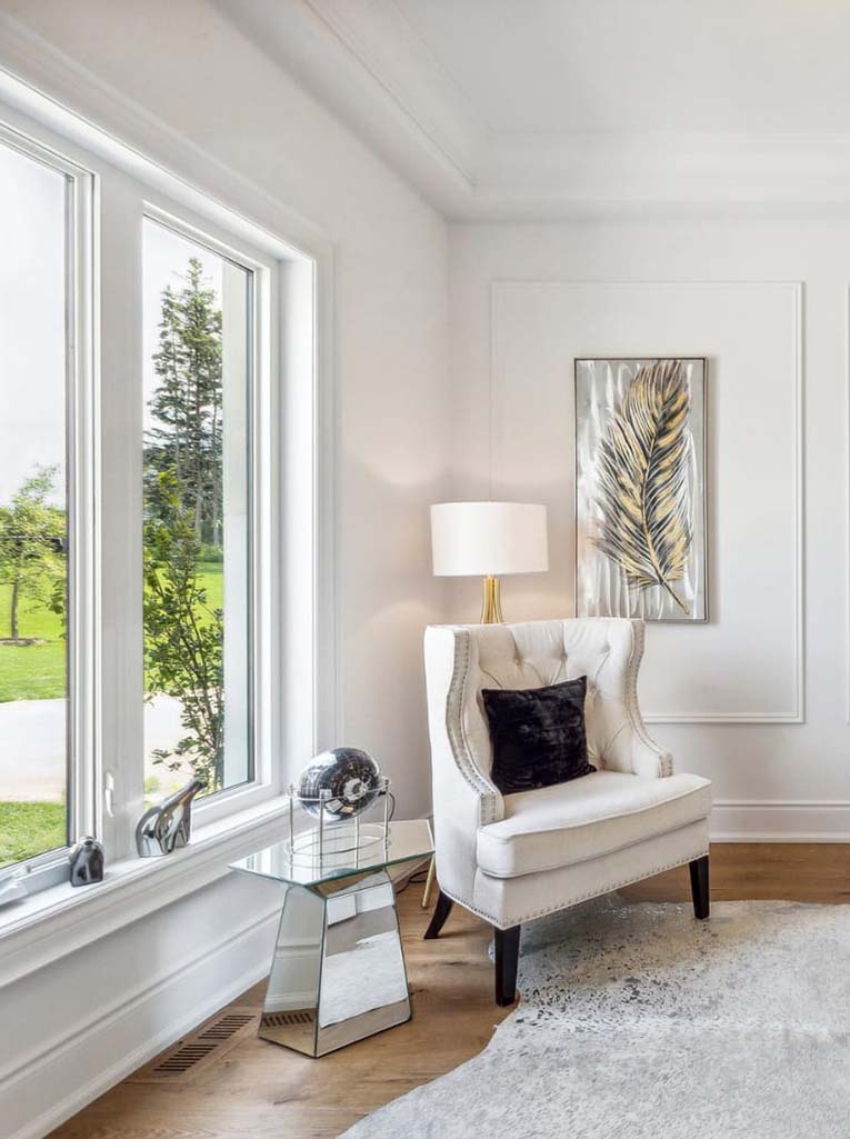 Sitting room staged with white chair, dark pillow and floor lamp. Nice window with nature outside.